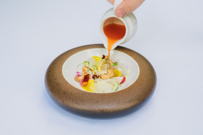 Hand pouring sauce into round plate filled with vegetables, fruit and edible flowers