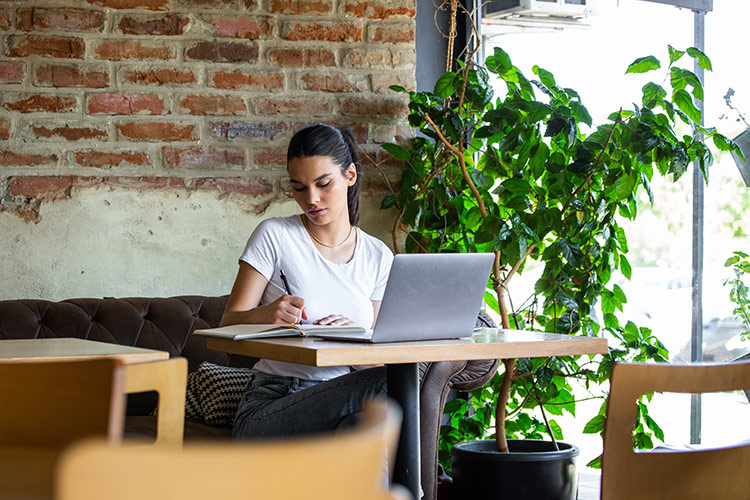 a woman sitting on the bench working at a laptop and writing in her book