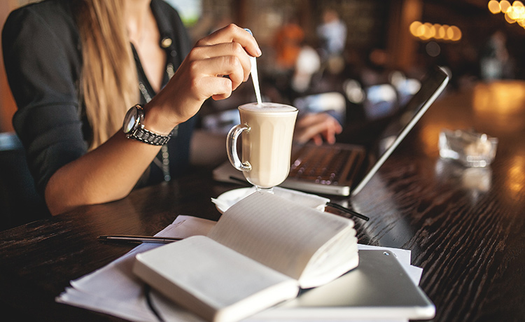a woman stirring her coffee with a laptop and book in front of her